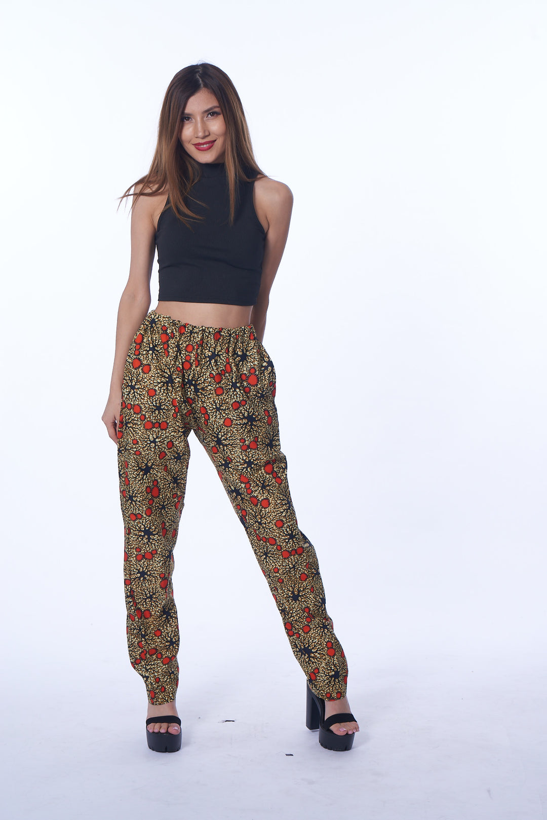 A photo of a woman wearing myObioma’s Ameena Pants. These beige harem pants feature an elastic waist and ankle design that keeps the pants in position above the waist. The wide leg feature emphasizes the legs, especially with the brown and orange circle patterns scattered throughout the print.