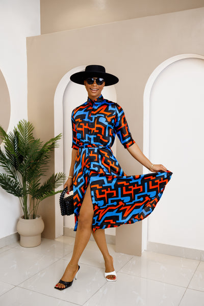 Woman wearing a vibrant Akwu shirt dress with a bold blue, black, and orange geometric print, accessorized with a black hat, sunglasses, and heels.