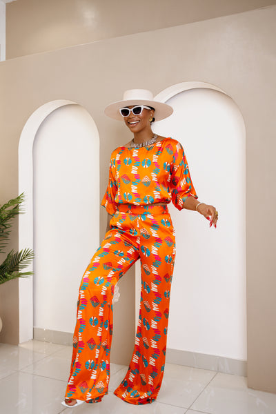 Woman wearing a vibrant orange wide-leg pant set with African-inspired prints, styled with a white hat, sunglasses, and statement accessories.