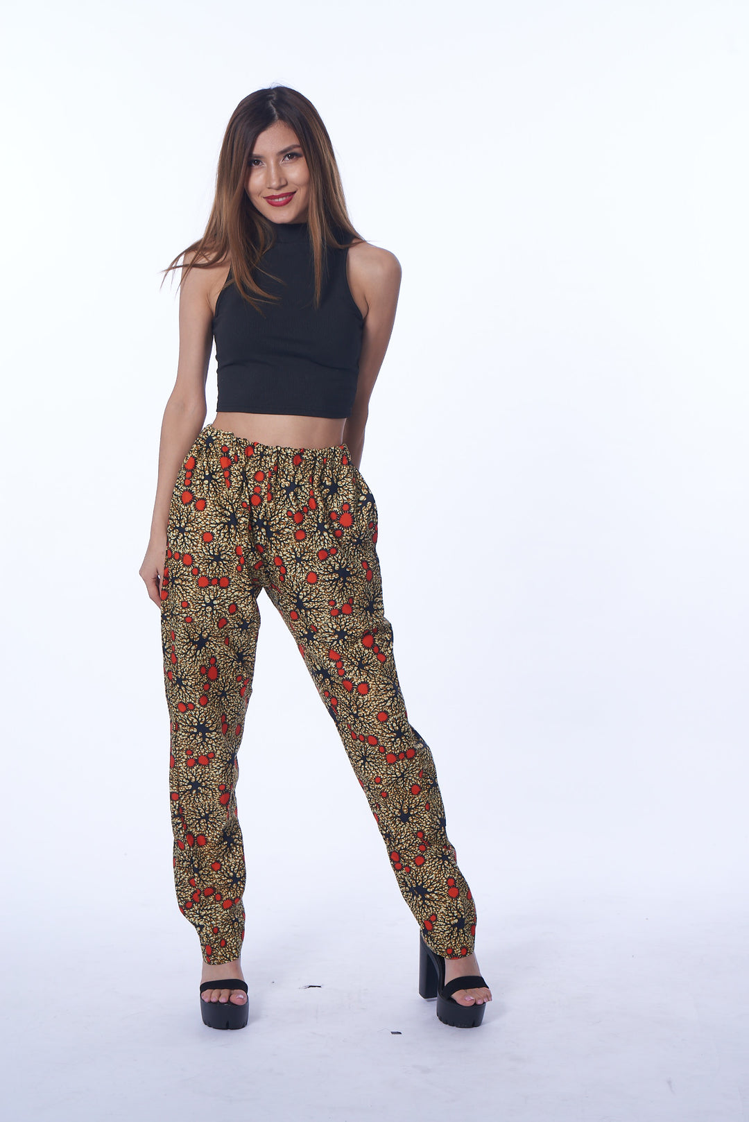  A photo of a woman wearing myObioma’s Ameena Pants. These beige harem pants feature an elastic waist and ankle design that keeps the pants in position above the waist. The wide leg feature emphasizes the legs, especially with the brown and orange circle patterns scattered throughout the print.