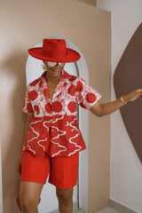 Person wearing a red and white patterned shirt and shorts with a matching hat, standing against a beige wall.