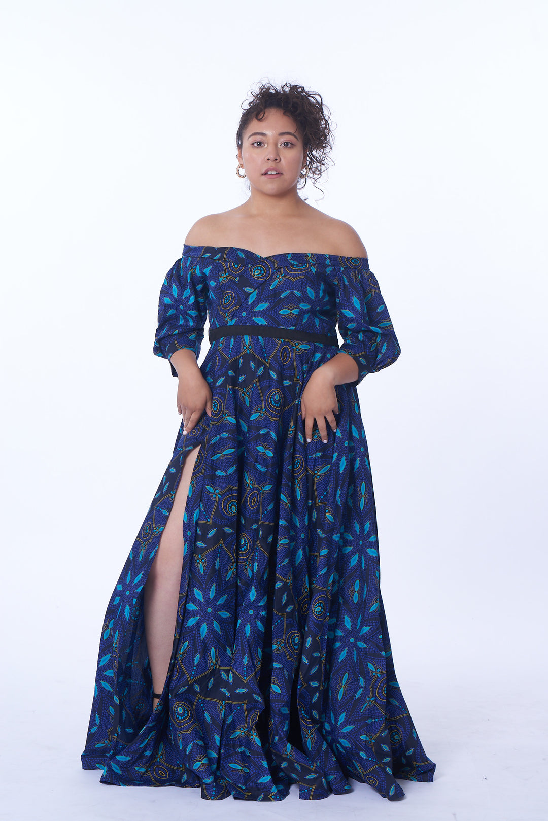 Photo of a woman wearing myObioma's long and blue Pokou Dress. The dress includes a slit thigh and off-shoulder puffy sleeves, while being filled with deep blue patterns and golden accents. It also includes a thin, black belt around the waist.