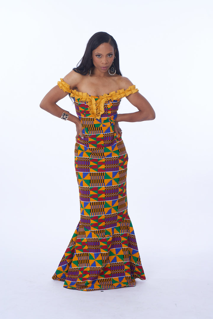 Photo of a woman wearing myObioma's Akua Kente Dress, which is long and multicolored. The dress includes yellow, ruffled off-shoulder straps and vibrant geometric patterns. The attractive fitted bodice includes a hem that flares out for a drastic flair.