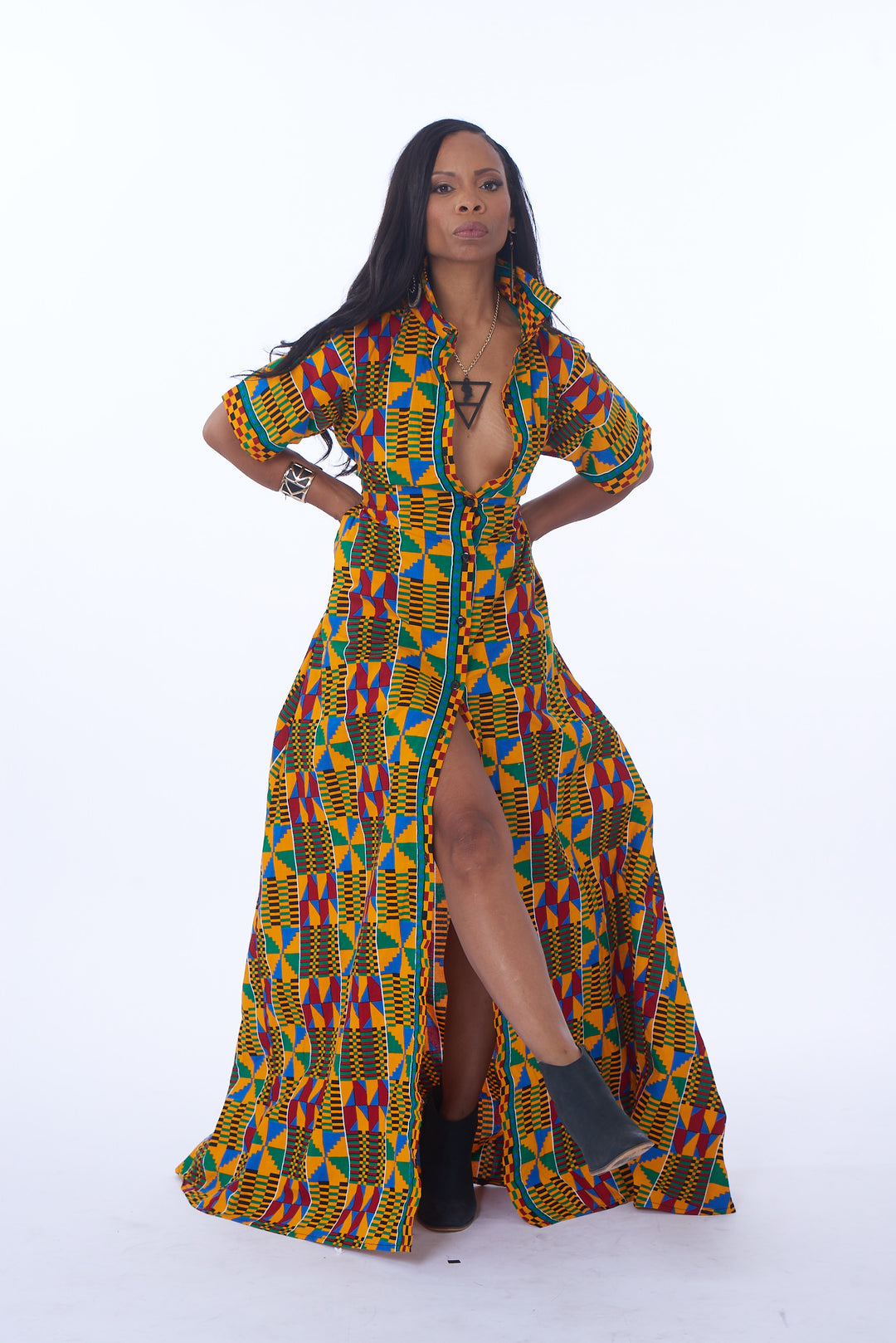 Photo of a woman wearing myObioma's Mawusi Kente Wrap Dress, which is long and printed with multicolor geometric patterns. It includes a button-up feature, medium-length loose sleeves, and a collar for dramatic flair. 
