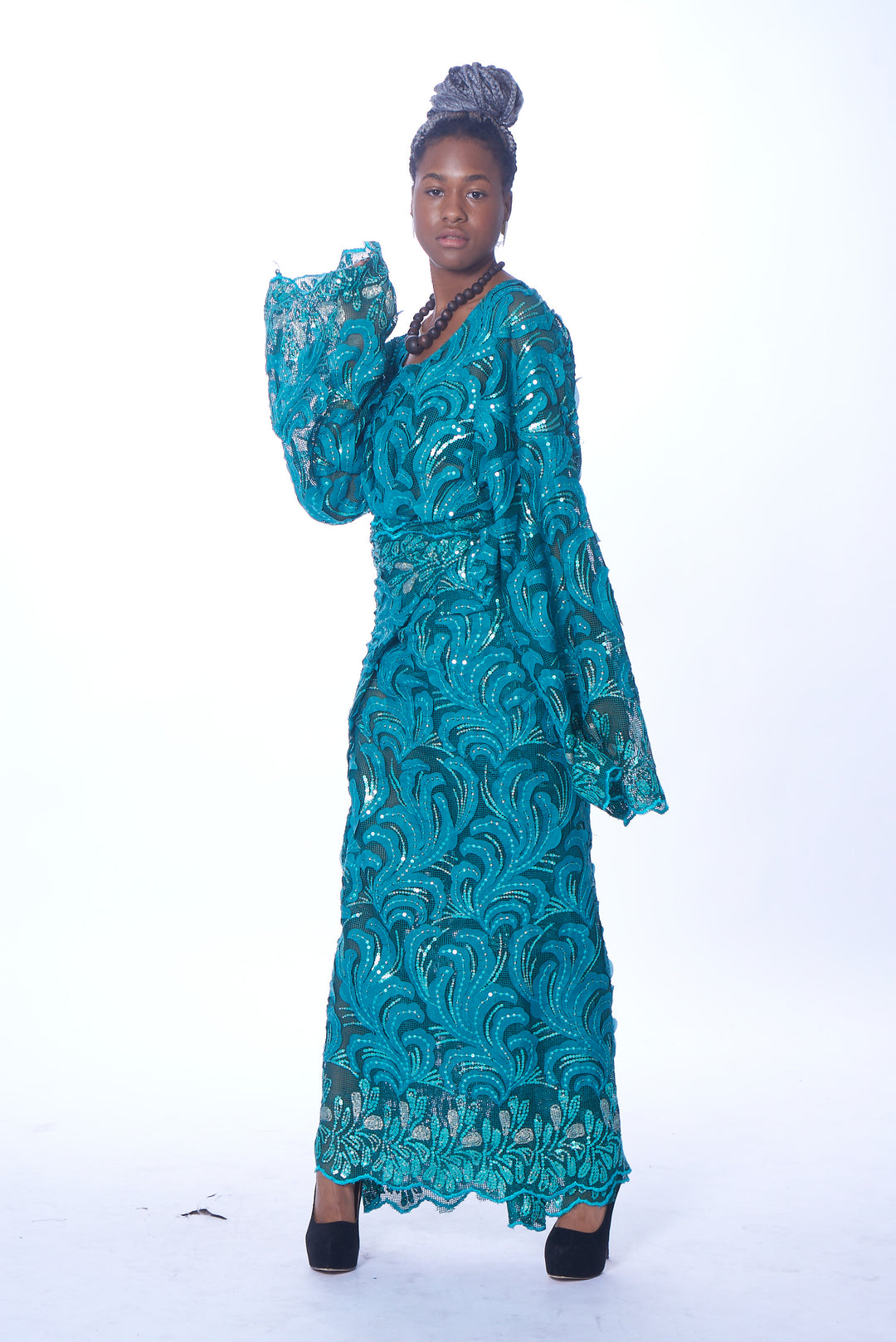 Photo of a woman wearing myObioma’s Chinenye Trad Dress, which is a column maxi aquamarine dress. The modest and feminine dress features flared long sleeves and a v-neck for an elegant appearance. The sheer lace fabric throughout the dress is adorned with embroidered curvy patterns. It also has a matching, lacy belt to emphasize the waist. 