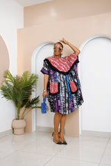 Model wearing Nwayiocha Adire Midi Dress in black, purple, and blue stripes with red patterned yoke and pockets trimmed with black fringe, holding blue beaded bag.