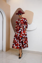 Woman wearing the Nwayioma Wrap Dress, a voluminous red African print wrap dress with pockets, styled with a wide-brim hat, sunglasses, and black heels.