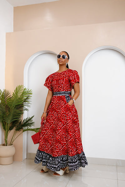 Model wearing red Obataobie Ankara Maxi Dress with flutter sleeves and black-and-white hem, standing confidently in a minimalist indoor space.