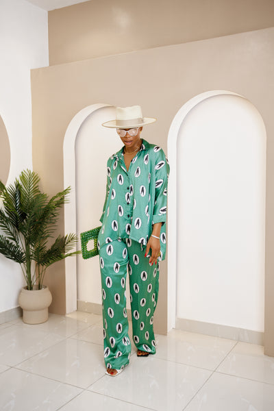 Woman wearing a stylish green silk co-ord set with a modern African print, oversized button-down shirt, and wide-leg pants, accessorized with a hat and bag.