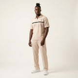 Model wearing the Okwuru beige men’s knit two-piece set with a short-sleeve zip polo, geometric detailing and relaxed straight-cut drawstring pants.