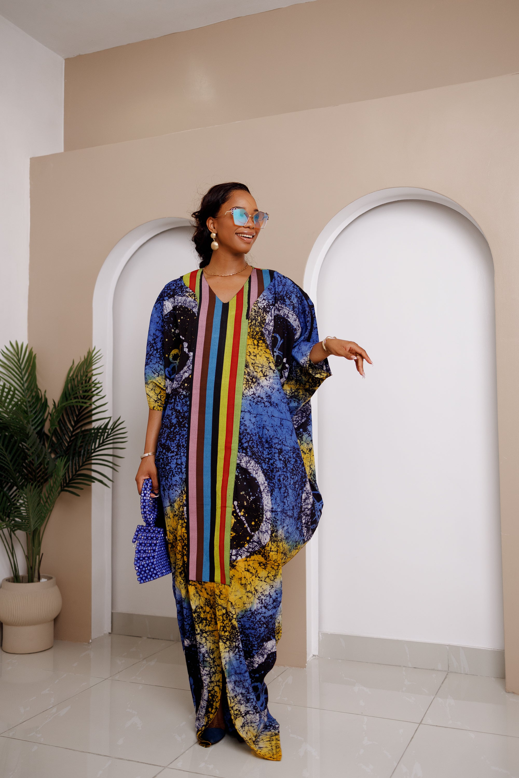 Woman wearing Onwenkem Adire Bubu dress with colorful Adire print and bold Aso Oke panel down the front, styled with blue sunglasses and clutch.