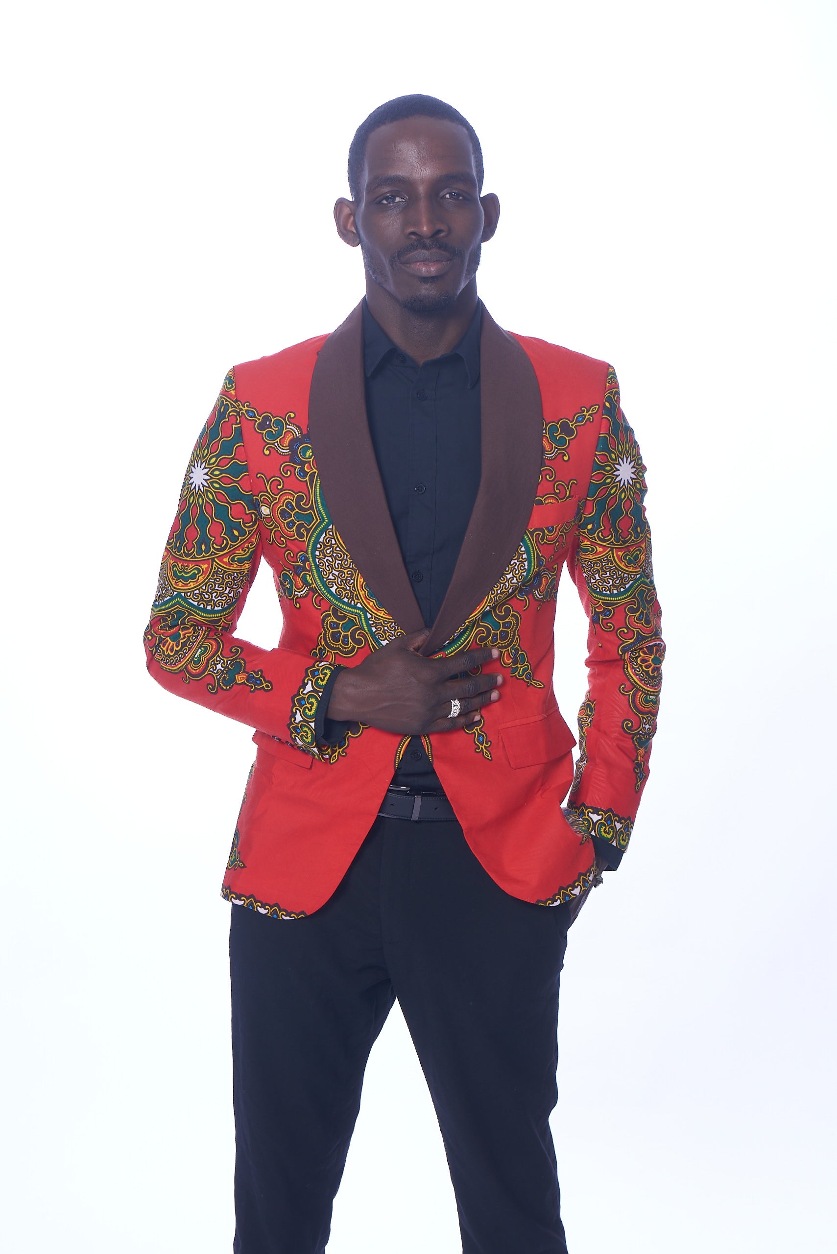 Photo of a man wearing myObioma's red Dashiki Blazer. It features beautiful green and yellow geometric patterns that loop around the sleeves and dark lapels. Classic red flap pockets are included on each side for usage and subtlety as they blend perfectly into the blazer. 