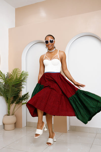 Woman wearing a white top and red and green skirt in a room with a plant and beige wall.
