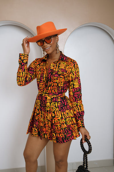 Woman wearing a colorful patterned outfit with an orange hat and sunglasses, standing against a white wall.