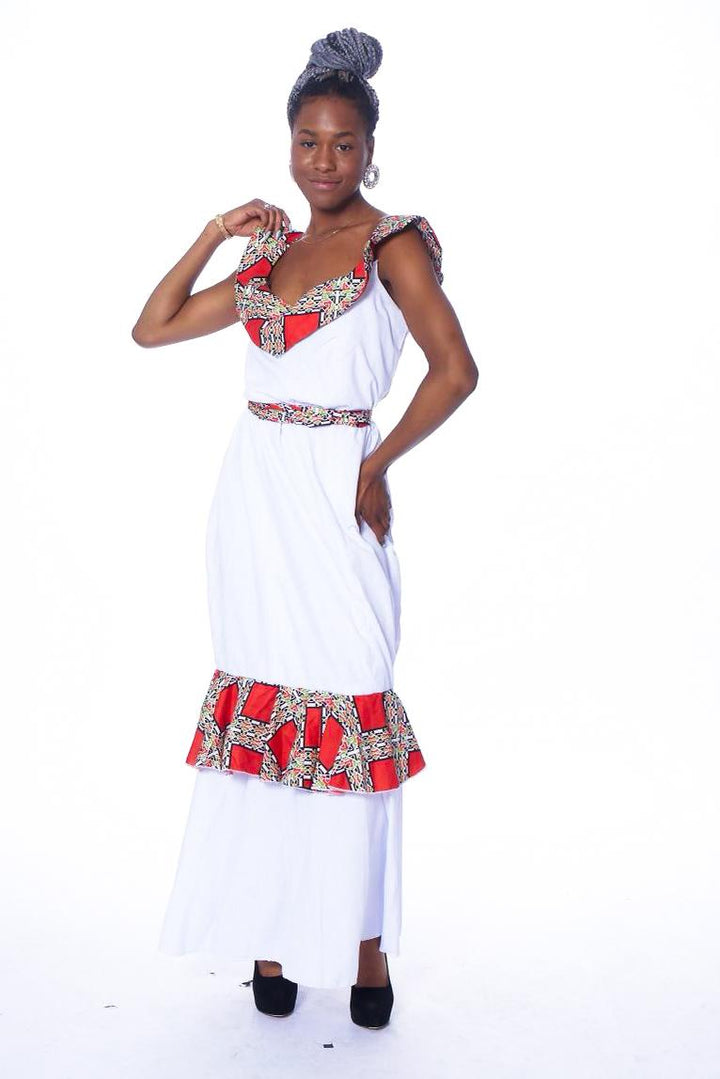 Photo of a woman wearing myObioma’s Nonye dress. The long white dress includes a belt around the waist and a ruffled feature around the knees. It also has a v-neck design that exposes the neck and upper chest, connecting to the dress with sleeveless shoulder straps. The shoulder straps, belt, and knee ruffle feature a red pattern design that stands out against the white fabric. 
