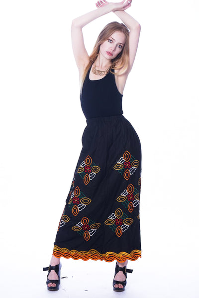 Photo of a woman wearing myObioma's Makda Ethiopian Skirt, which is long and black. The A-line skirt includes scattered multicolor flower-like embroidered patterns down to the hem.