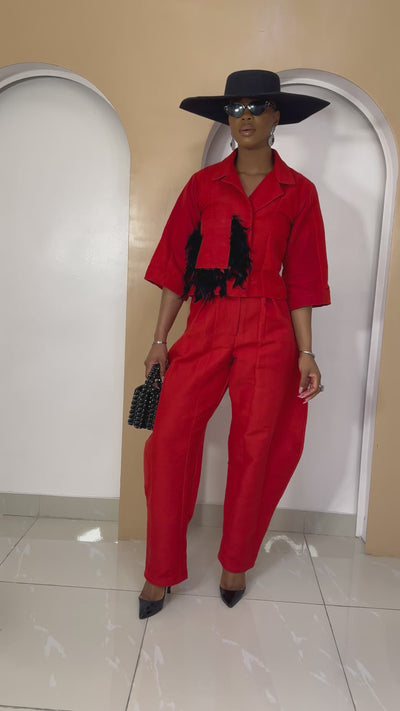 Female wearing Nnekota Aso Oke red two-piece set with wide-leg trousers and a structured top decorated with black feather trimmings.