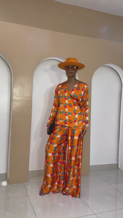 A model wearing a bright orange Ankara silk wrap top with multi-colored patterns, paired with matching wide-leg palazzo pants, styled with a bold orange hat.