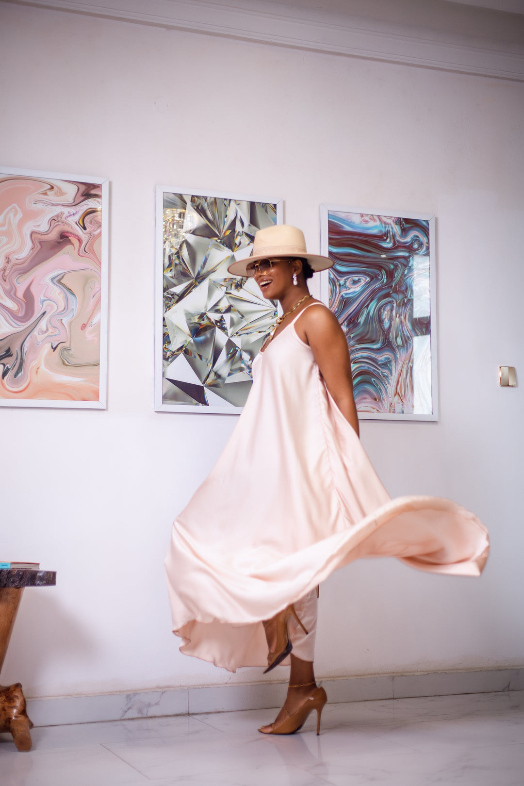 A woman posing in a cream silk maxi dress. She is pictured spinning around and laughing. She is styled wearing a tan sun hat, gold jewelry, and nude high heels.