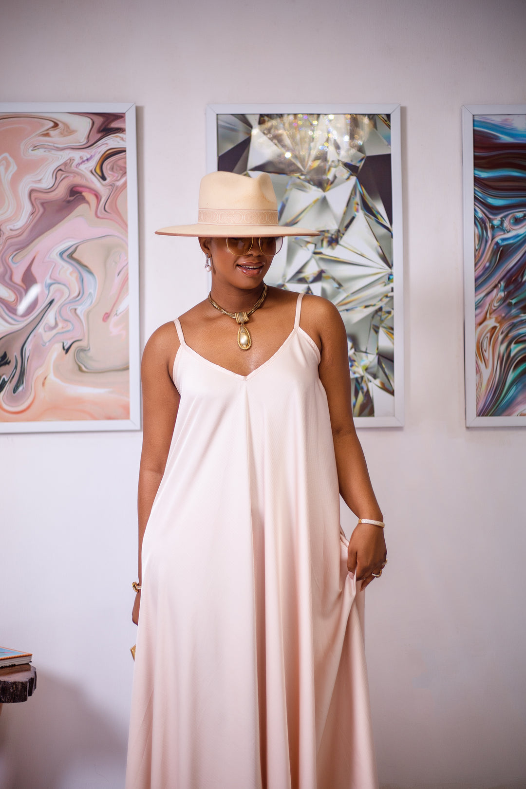 A woman posing in a cream silk maxi dress. She is pictured facing the camera and smiling. She is styled wearing a tan sun hat and gold jewelry. 