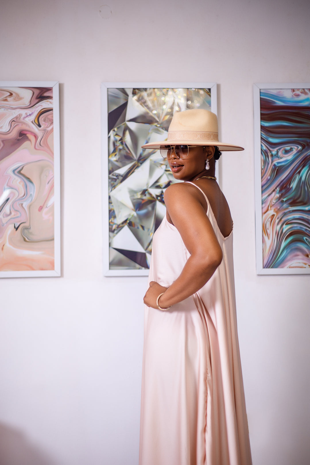 A woman posing in a cream silk maxi dress. She is pictured with her hand on her hip, facing to the side, and looking back at the camera. She is styled wearing a tan sun hat and gold jewelry.