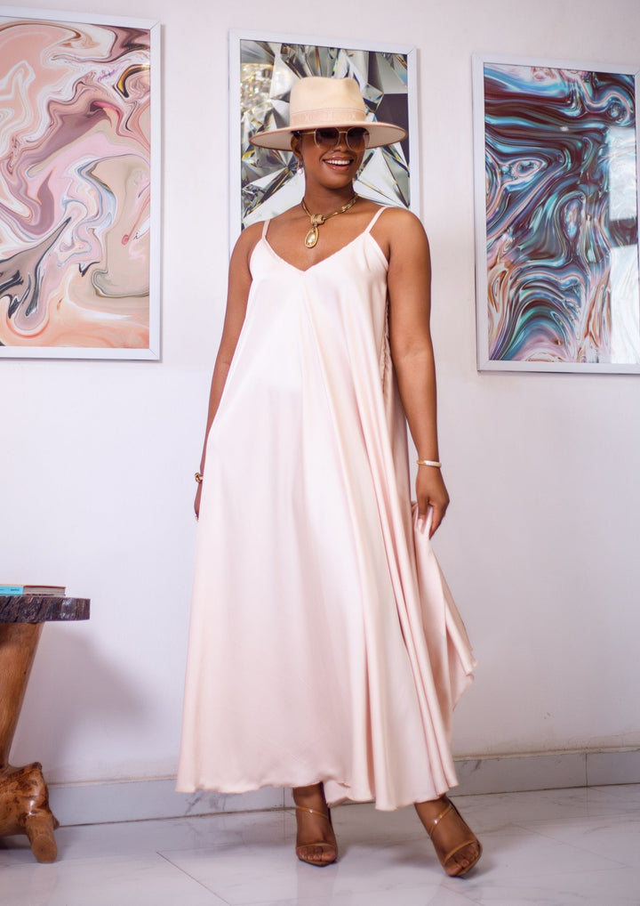 A woman posing in a cream silk maxi dress. She is pictured facing the camera with her arms at her sides. She is styled wearing a tan sun hat, gold jewelry, and nude high heels.