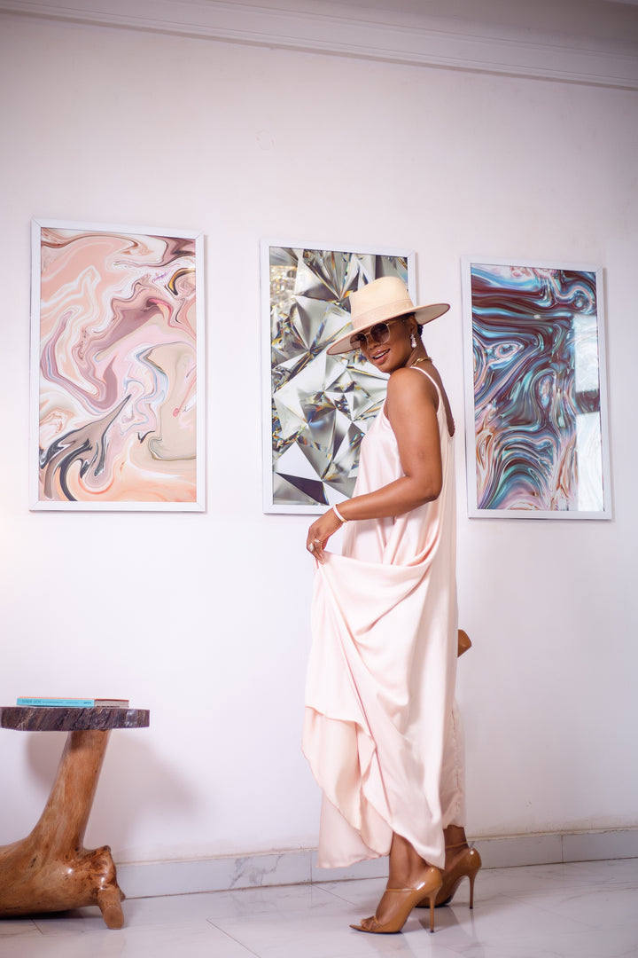 A woman posing in a cream silk maxi dress. She is pictured turning to the side and looking over her shoulder. She is styled wearing a tan sun hat, gold jewelry, and nude high heels.