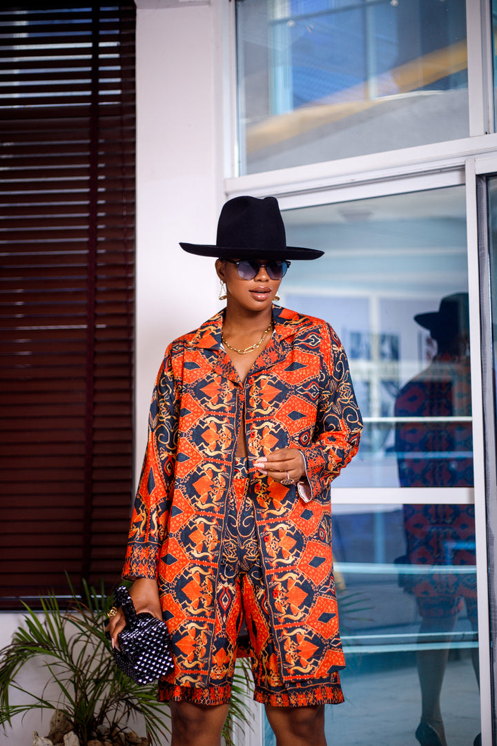 A women posing in a navy blue and burnt orange African print shirt and shorts two piece set. She is shown facing the camera. She is styled wearing a black sun hat, gold jewelry, and a black hand bag.
