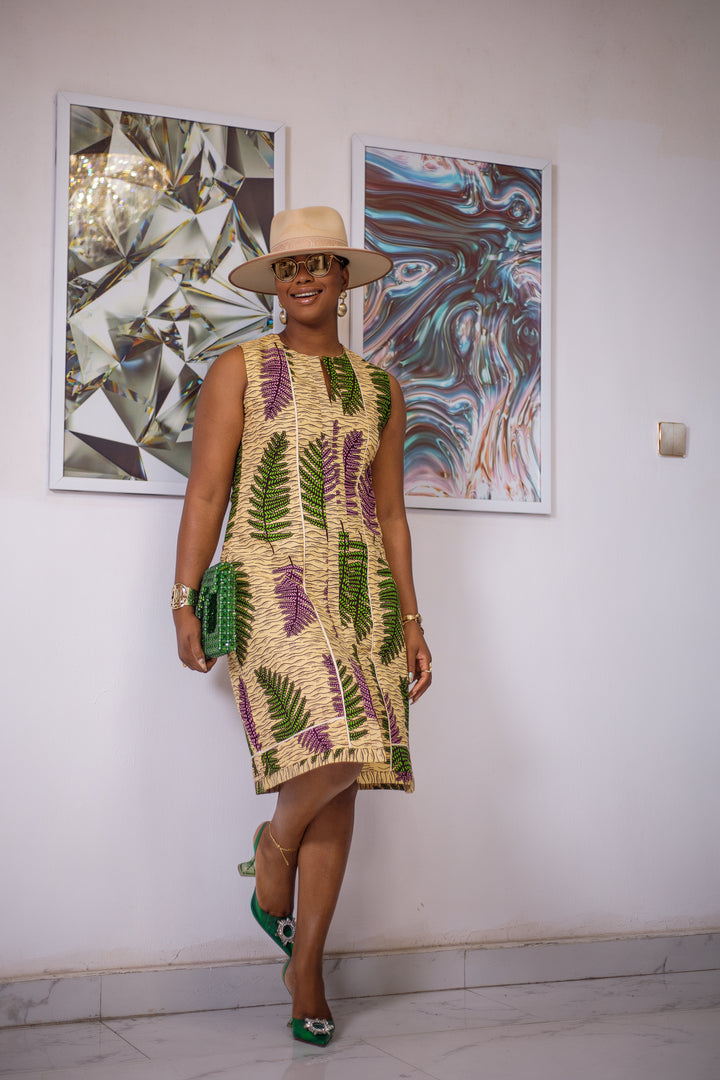 A woman posing in a purple and green African print midi dress. She is styled with a tan sun hat and a green purse.
