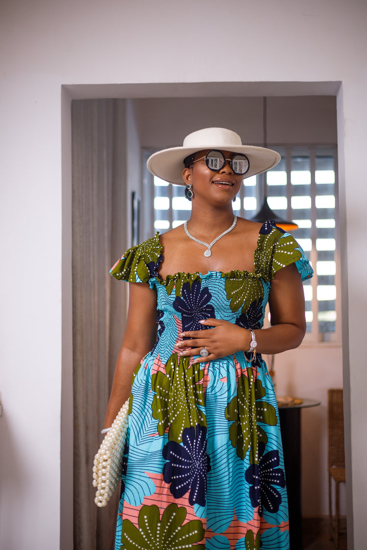 A woman posing in a blue and green African print mid-length dress. She is facing the camera and smiling. She is wearing a white sun hat and silver jewelry.