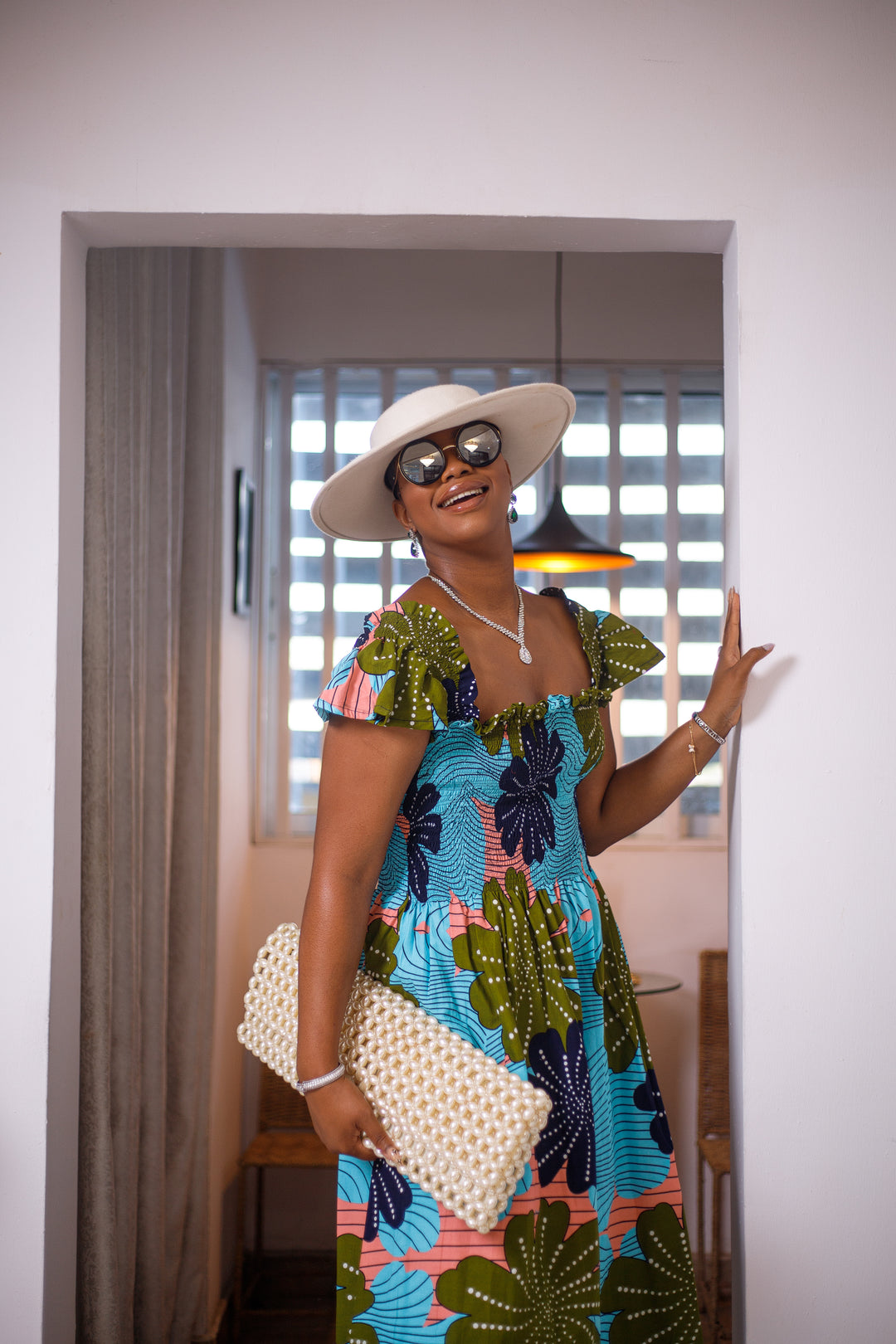 A woman posing in a blue and green African print mid-length dress. She is facing the camera and smiling. She is wearing a white sun hat and silver jewelry.