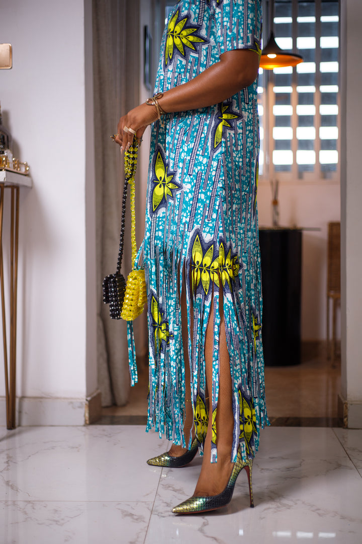 A woman posing in a blue, green, and white maxi dress with frills. She is pictured from the waist down. She is styled with a yellow and black beaded purse and high heels.