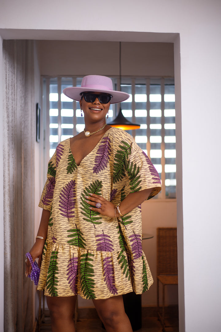 A woman posing in a mid length green purple and tan summer dress. She is pictured facing the camera with her left hand on her hip and smiling. She is styled with a purple hat and purse.