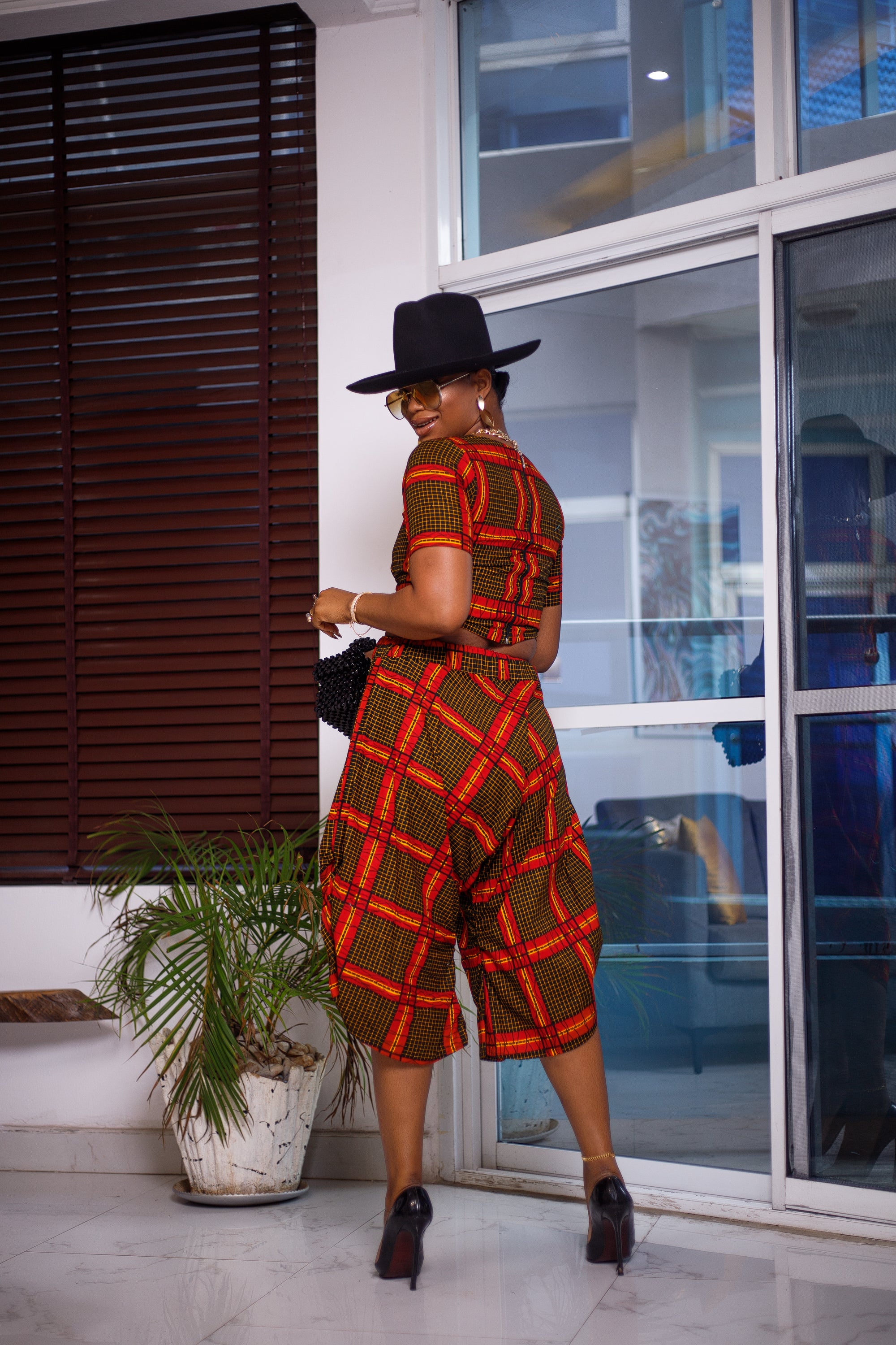 A woman posing in a yellow black and red two-piece set. She is pictured with her back to the camera and looking over her shoulder. She is styled with a black sun hat and silver jewelry.