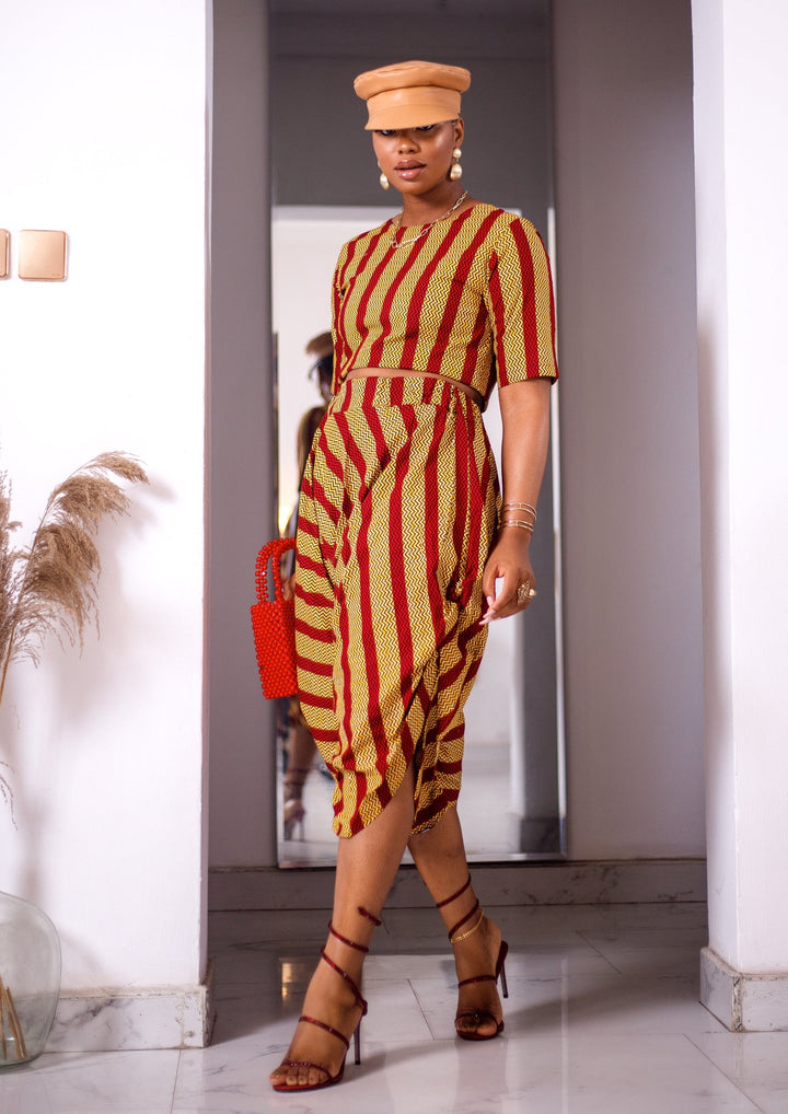 A woman posing in a yellow and red striped two-piece set. She is pictured facing the camera with her legs crossed. She is styled with a brown leather hat and a red beaded purse.