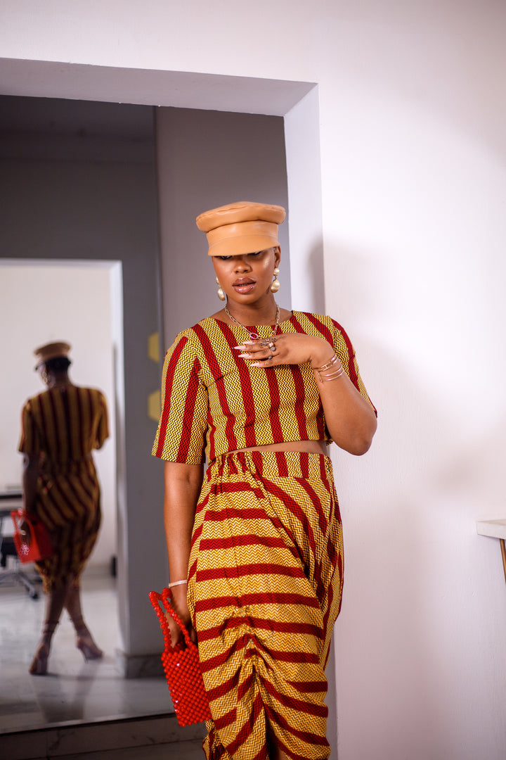 A woman posing in a yellow and red striped two-piece set. She is pictured facing the camera with her hand on her chest. She is styled with a brown leather hat and a red beaded purse.