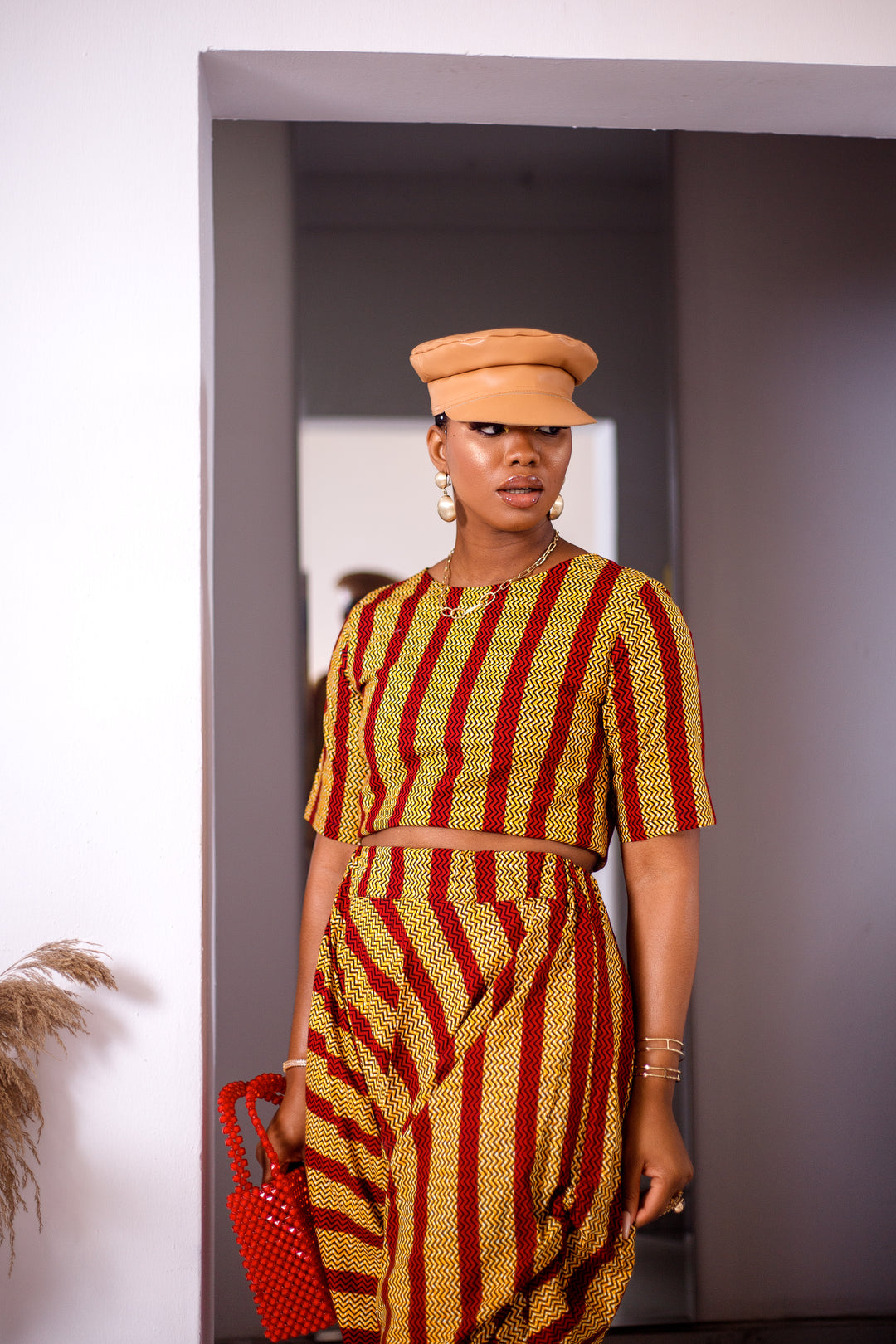 A woman posing in a yellow and red striped two-piece set. She is pictured facing the camera and looking to the side. She is styled with a brown leather hat and a red beaded purse.