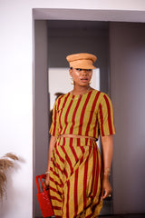 A woman posing in a yellow and red striped two-piece set. She is pictured facing the camera and looking to the side. She is styled with a brown leather hat and a red beaded purse.