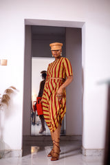 A woman posing in a yellow and red striped two-piece set. She is pictured facing the camera. She is styled with a brown leather hat and a red beaded purse.