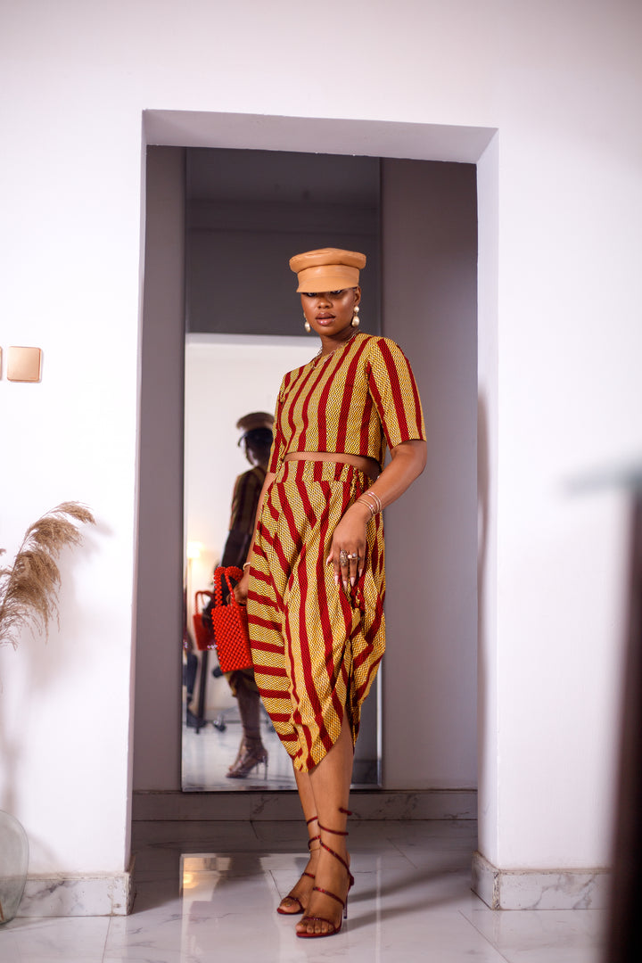 A woman posing in a yellow and red striped two-piece set. She is pictured facing the camera. She is styled with a brown leather hat and a red beaded purse.