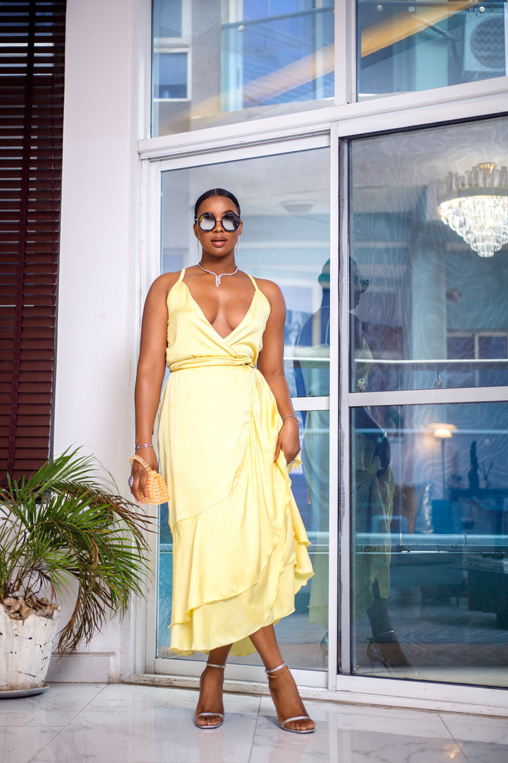 A woman posing in a pale yellow wrap around ruffle maxi dress. She is pictured facing the camera with her legs crossed. She is styled with brown sunglasses and silver jewelry.