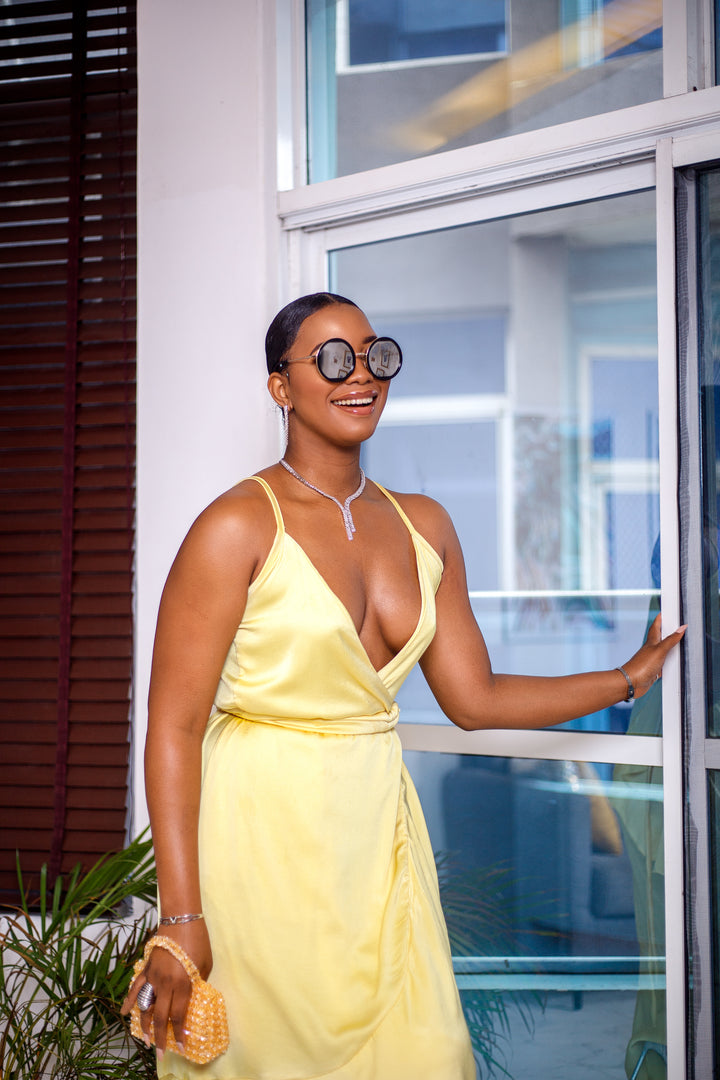 A woman posing in a pale yellow wrap around ruffle maxi dress. She is photographed from the waist up, turned to the side, and smiling. She is styled with brown sunglasses and silver jewelry.
