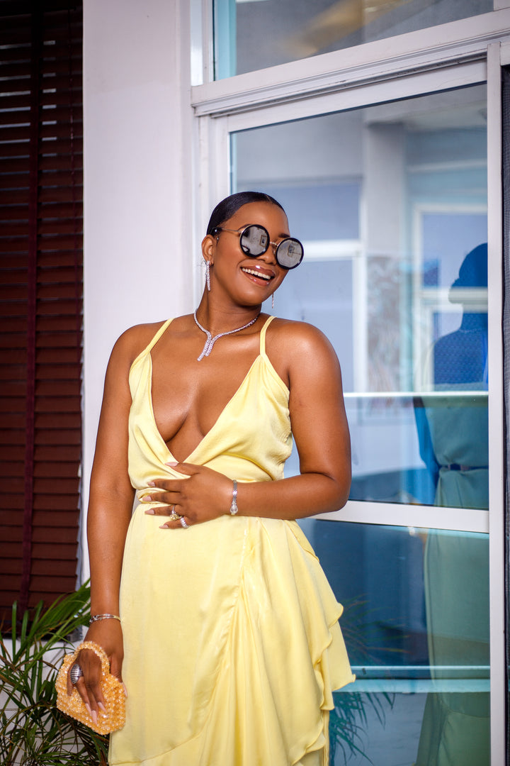 A woman posing in a pale yellow wrap around ruffle maxi dress. She is pictured from the waist up and smiling. She is styled with brown sunglasses and silver jewelry.