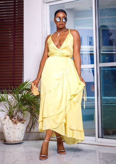 A woman posing in a pale yellow wrap around ruffle maxi dress. She is pictured facing the camera with her legs crossed. She is styled with brown sunglasses and silver jewelry.