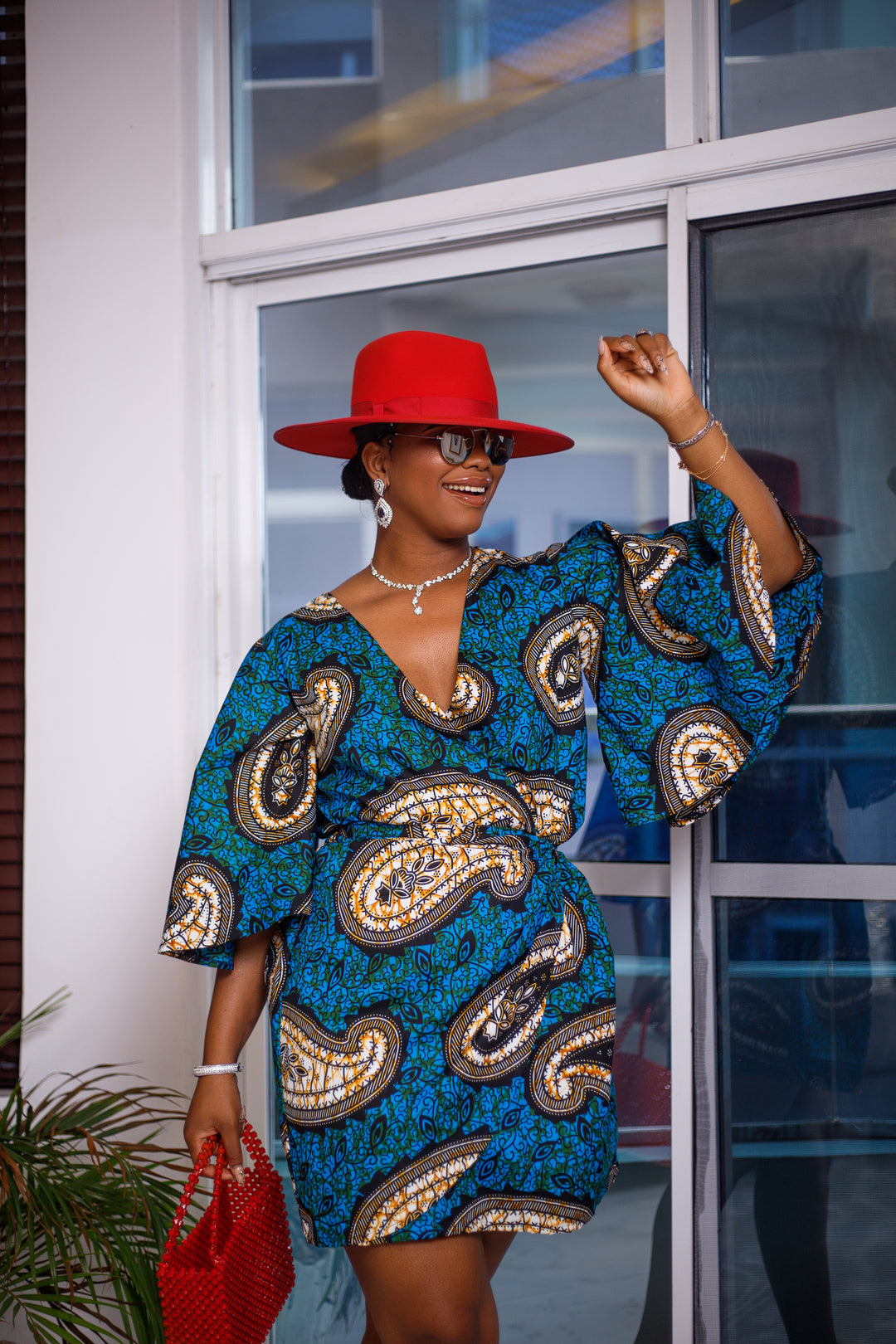 A woman posing in a blue and tan African print mini dress. She is pictured from the waist up and smiling. She is styled with a red beaded purse, a red sun hat, and silver jewelry.