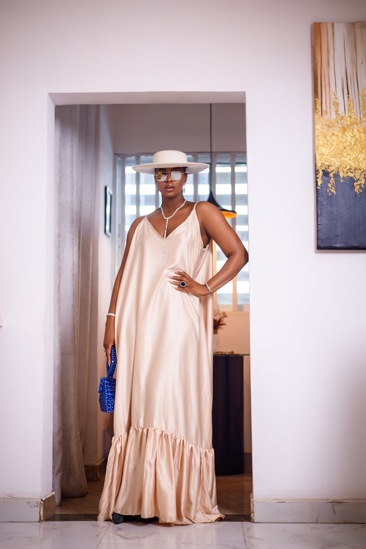 A woman posing in a pale pink silk maxi dress. She is pictured facing the camera with a hand on her hip. She is styled with a white sun hat, a blue beaded purse, and silver jewelry.