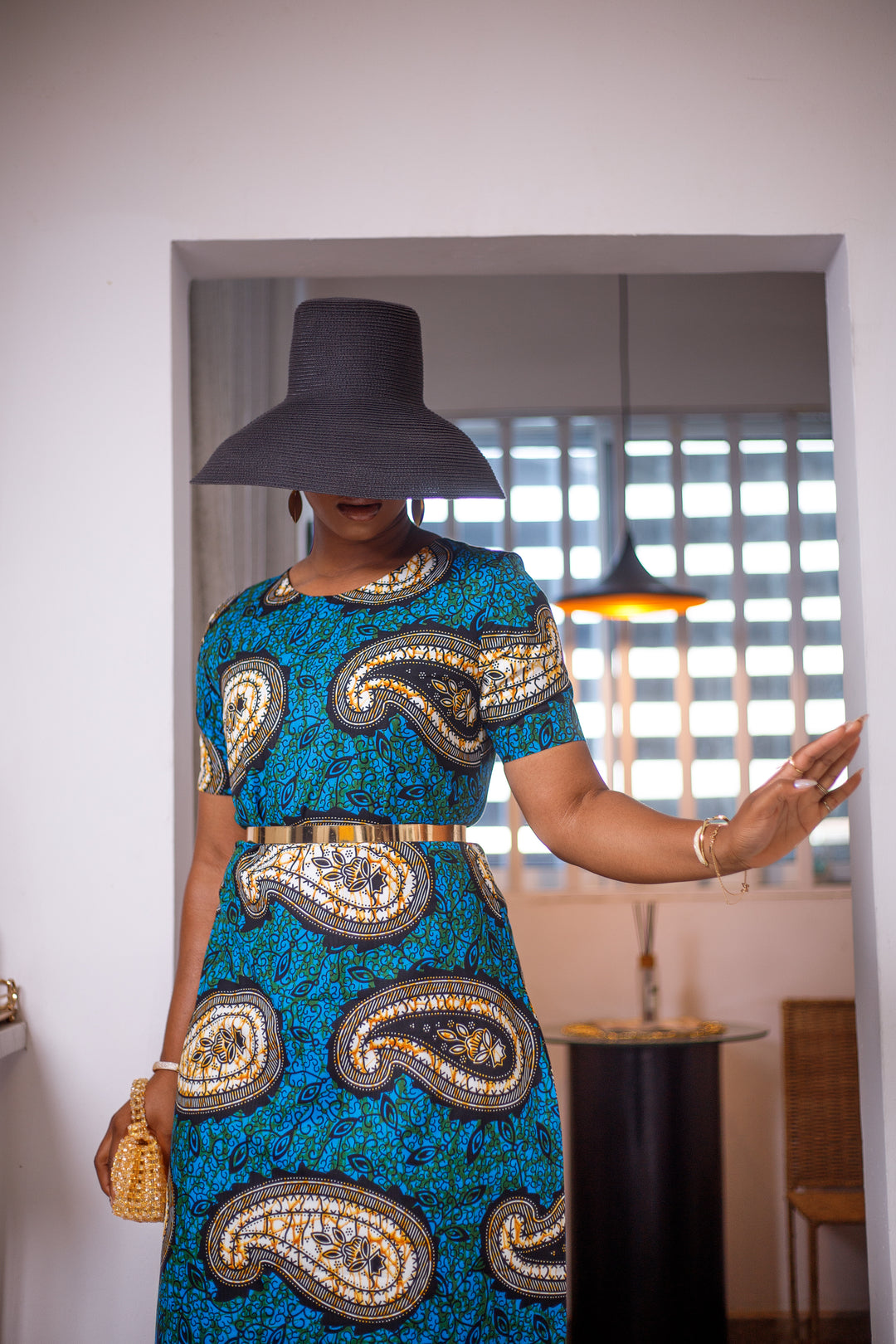 A woman posing in a black and blue African print maxi dress. She is styled with a black sun hat and a mini purse.