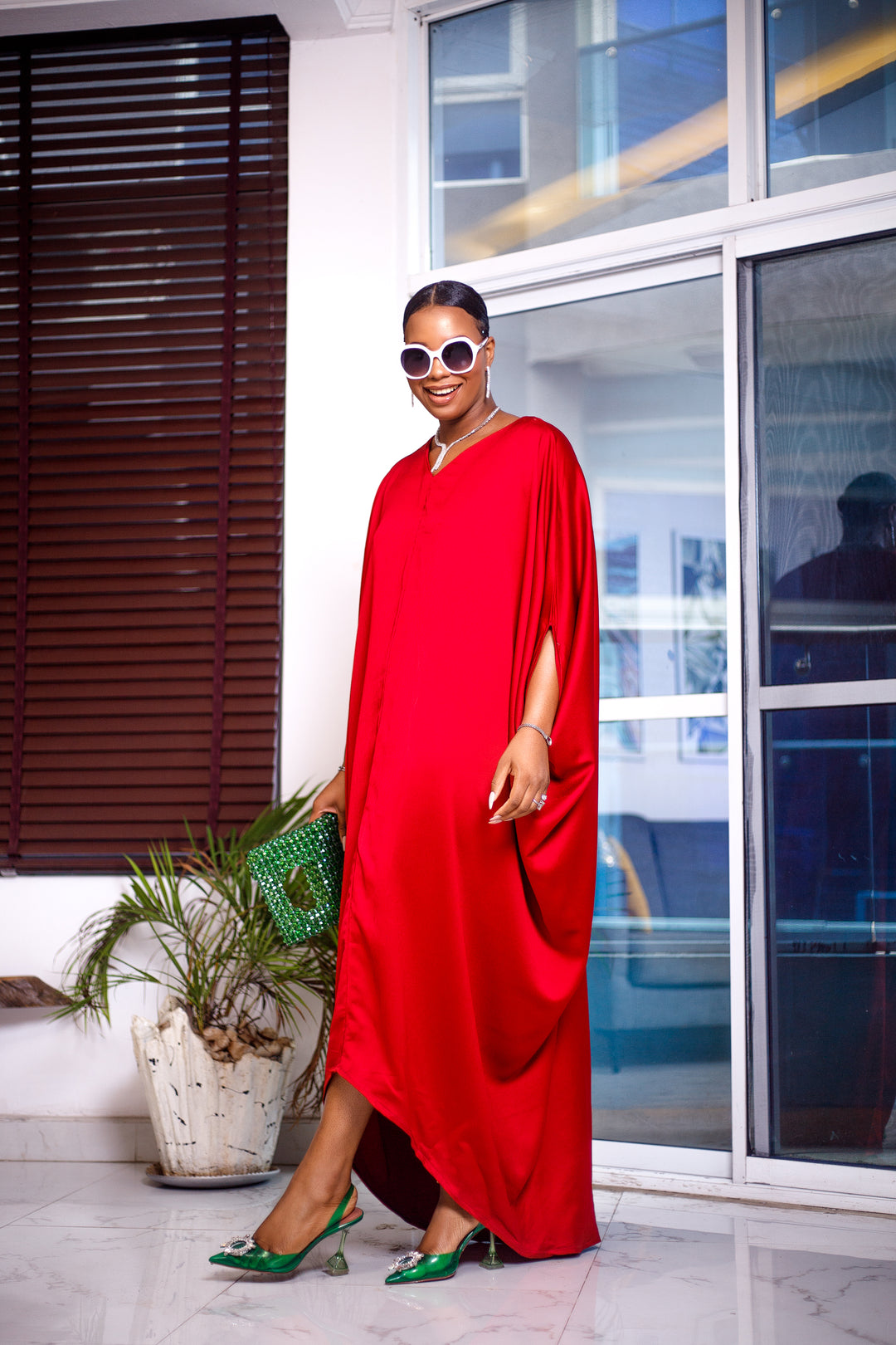A woman posing in a bright red maxi dress. She is pictured facing the camera and smiling. She is styled with white sunglasses and silver jewelry.