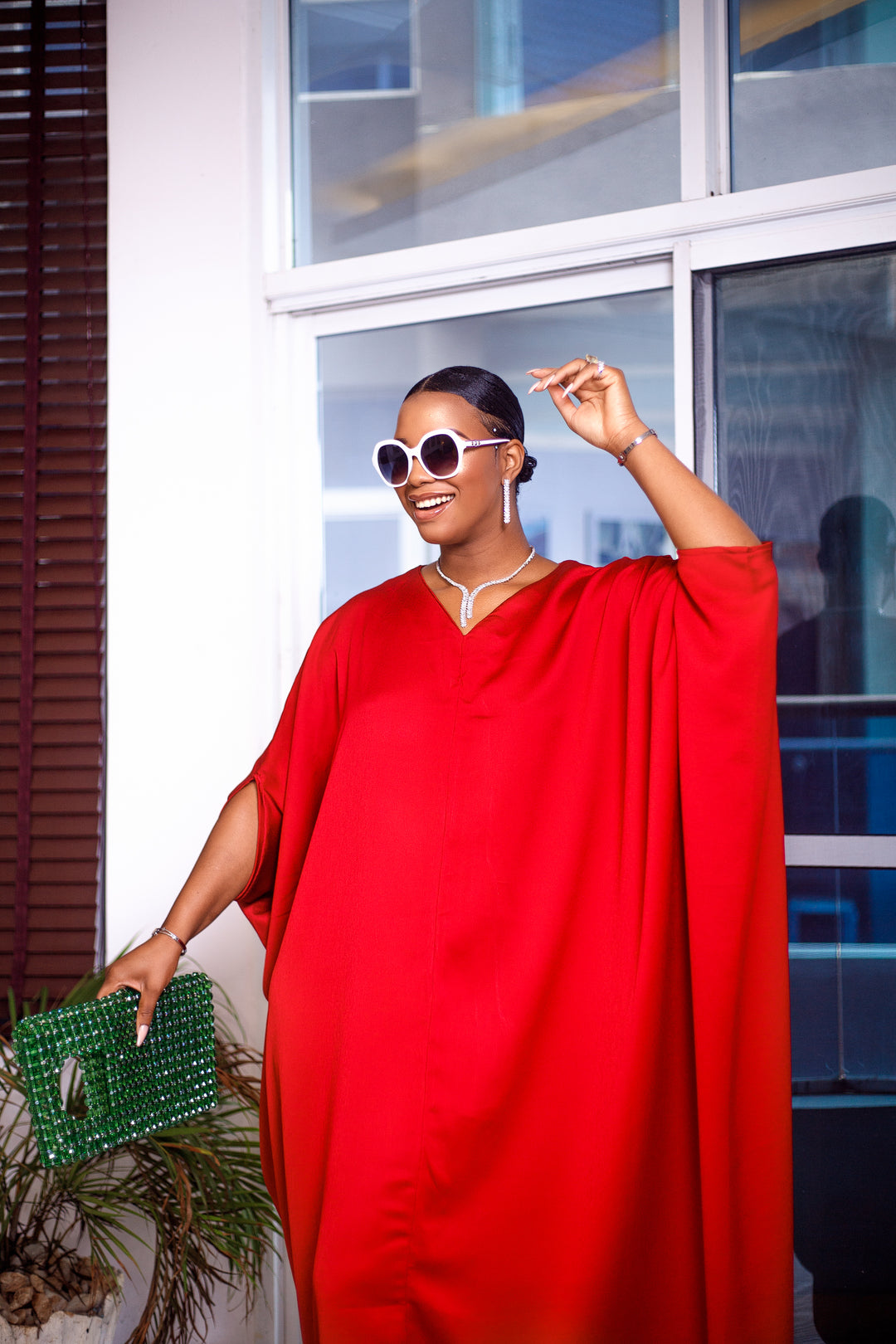 A woman posing in a bright red maxi dress. She is pictured from the waist up and smiling. She is styled with white sunglasses and silver jewelry.