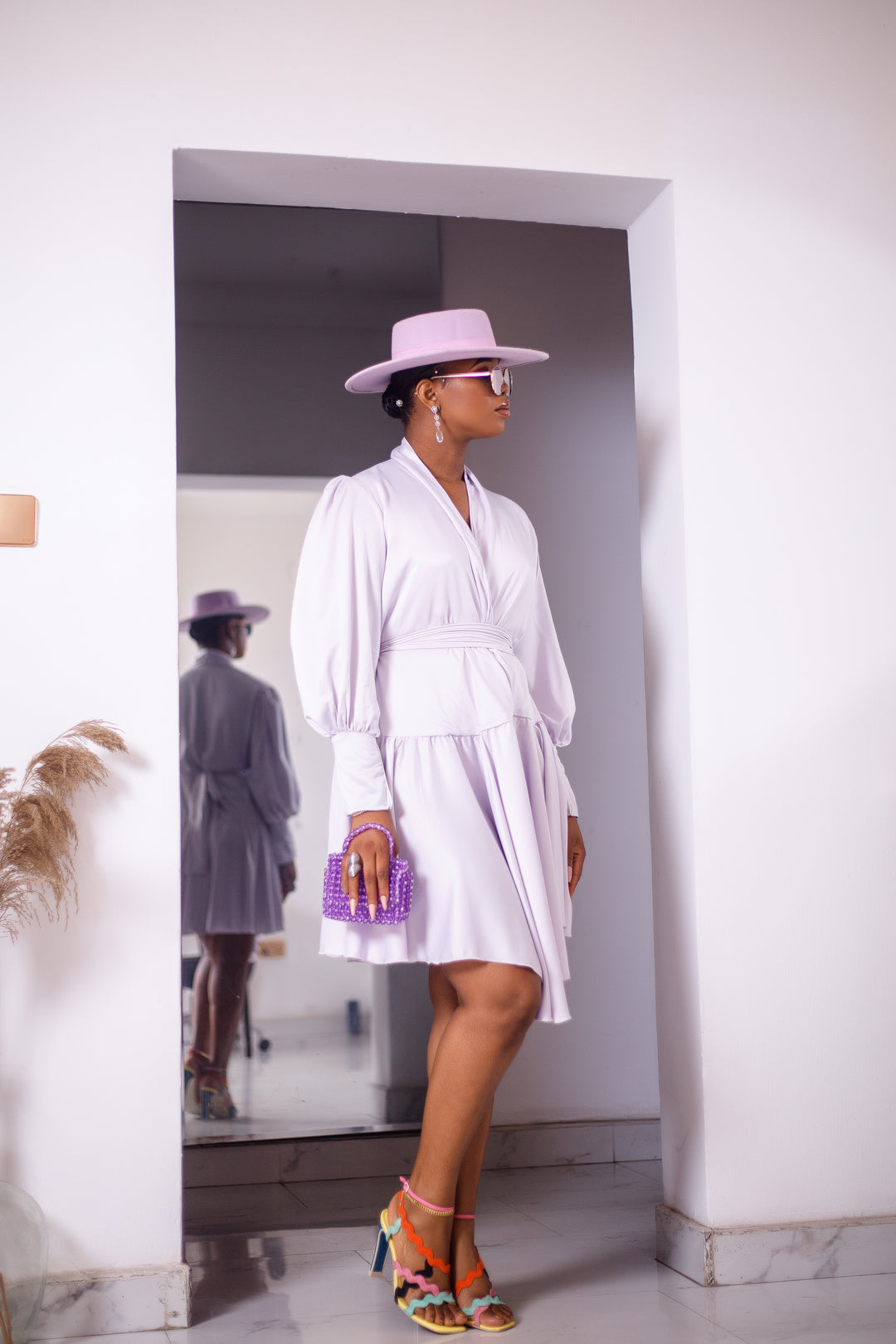 A woman posing in an unlined white silk dress. She is pictured facing the camera and looking off to the right. She is styled with a purple hat, a purple purse, and silver jewelry.